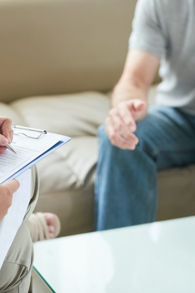 Faceless shot of anonymous woman sitting with patient during home visit and taking notes on clipboard